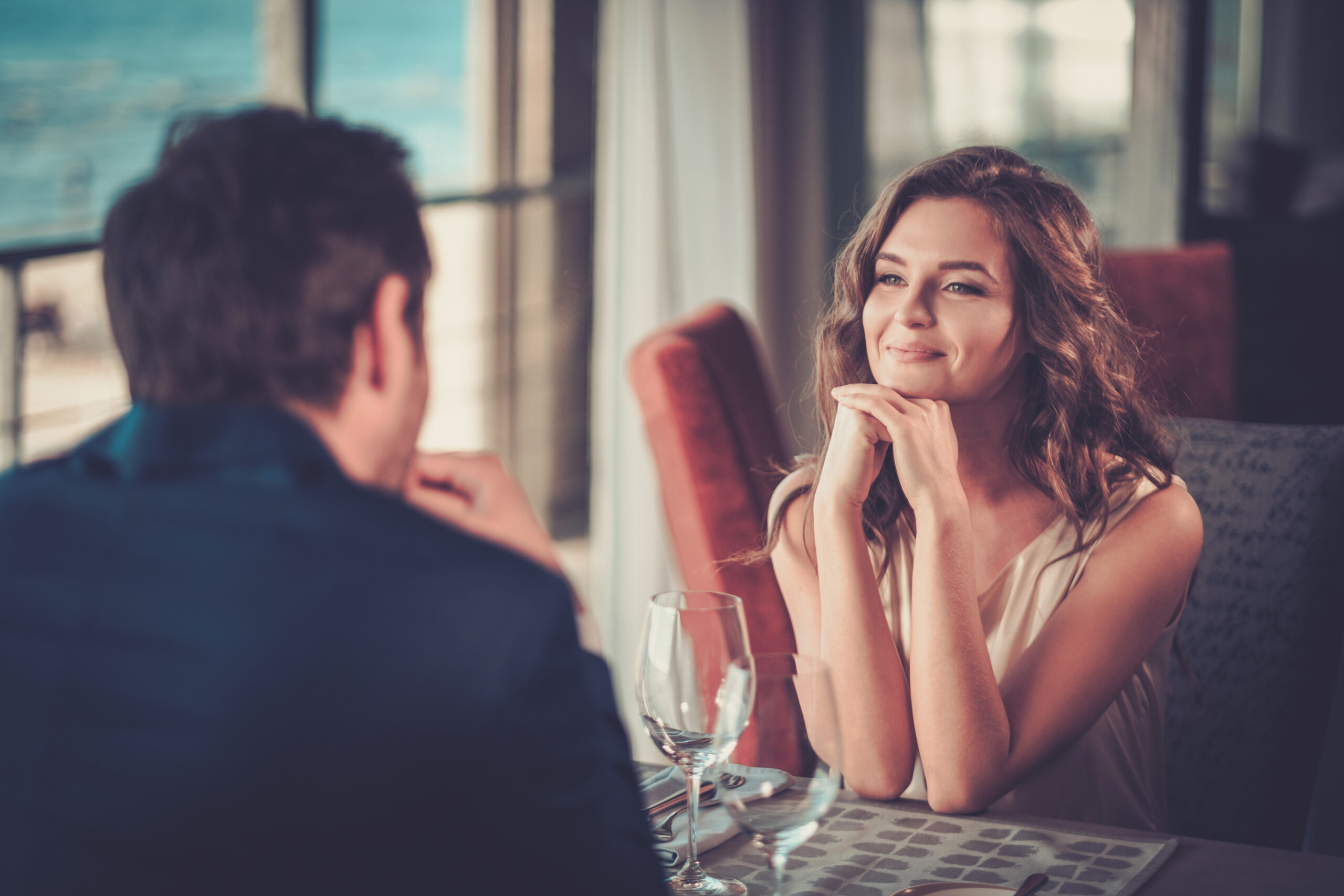 A smiling woman sitting across from a man at a restaurant table, resting her chin on her hands, with a serene ocean view visible through the window in the background.