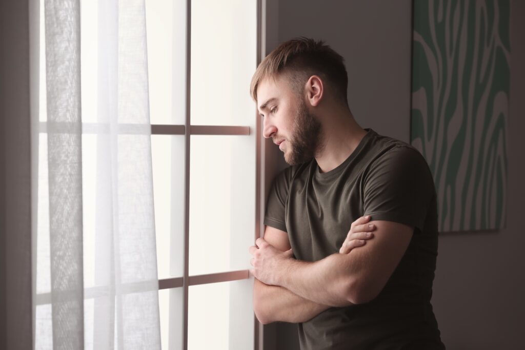 A young man with a beard and short hair stands by a window with his arms crossed, gazing outside with a contemplative expression.