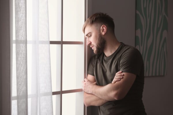 A young man with a beard and short hair stands by a window with his arms crossed, gazing outside with a contemplative expression.