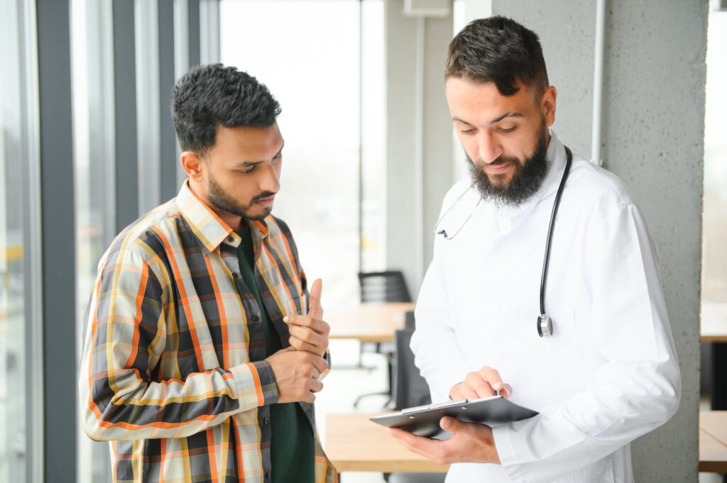 A doctor in a white coat with a stethoscope around his neck reviews a clipboard while speaking with a patient wearing a plaid shirt. The patient appears engaged, pointing at his chest as they discuss medical information in a bright office with large windows.