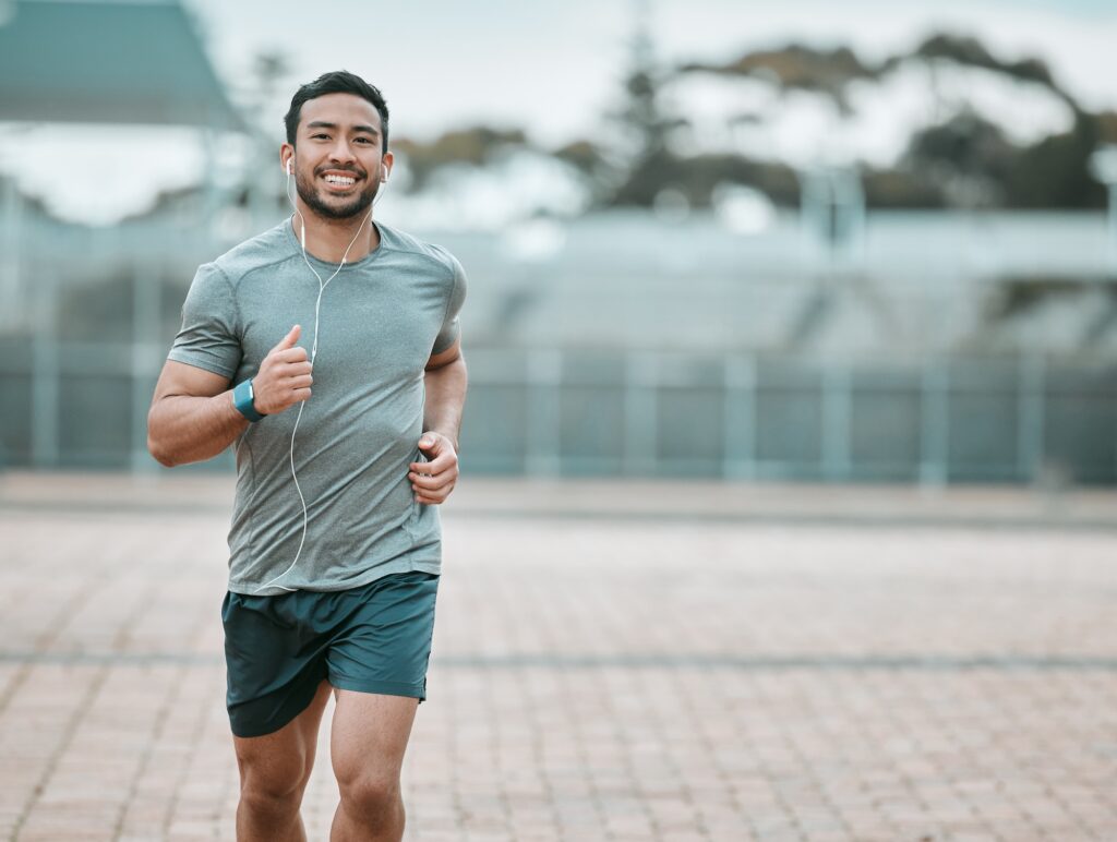 A young man in athletic wear runs outdoors with a smile on his face, listening to music through wired earphones. He wears a gray t-shirt, dark shorts and a fitness tracker on his wrist, with a blurred background of trees and a sports facility.