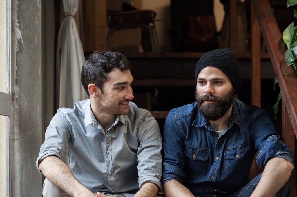 Two young men sit on a wooden staircase, engaged in conversation. One is wearing a light blue shirt and smiling, while the other, dressed in a denim jacket and black beanie, listens attentively.
