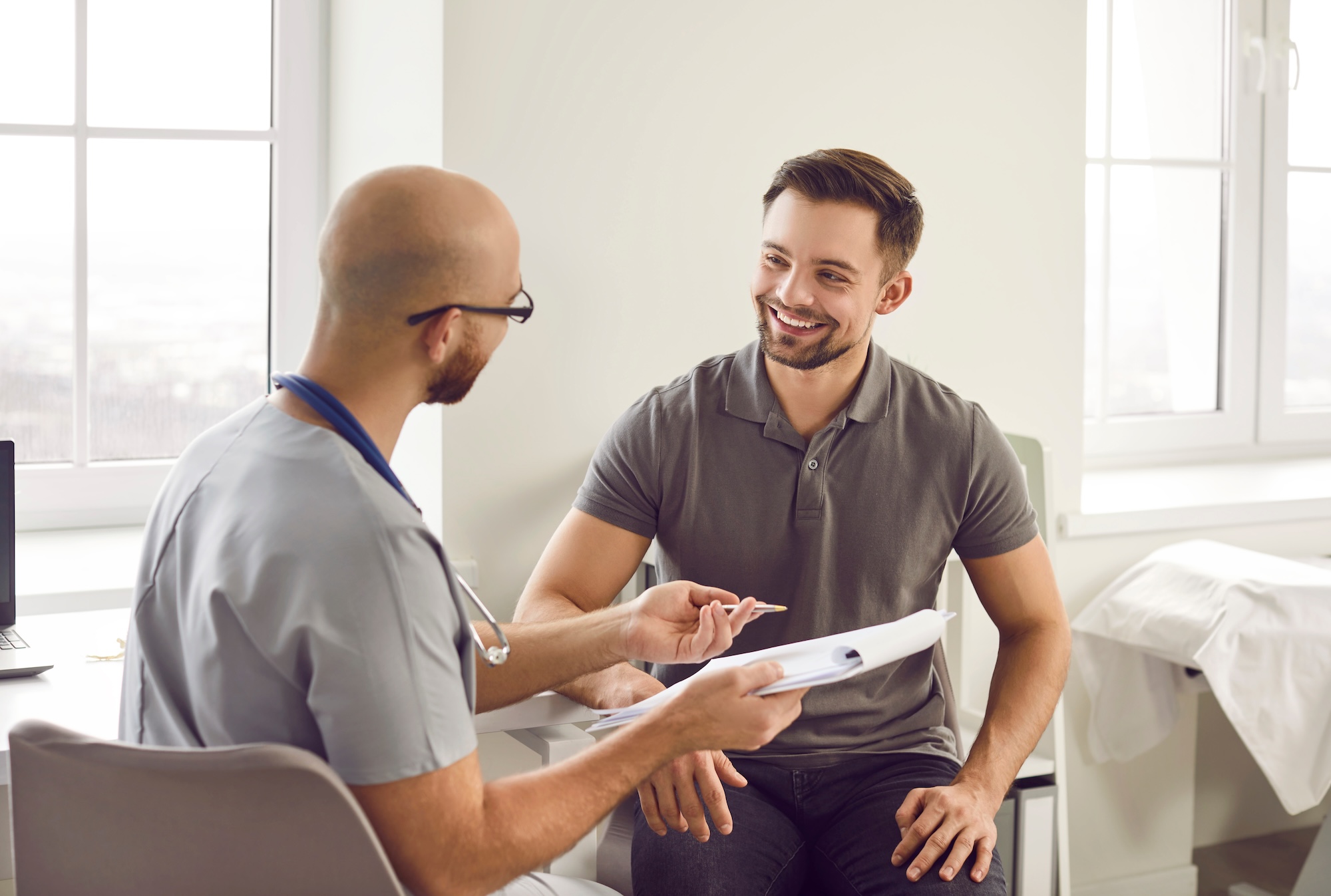 A doctor and a patient are sitting in a bright medical office. The doctor, wearing scrubs and a stethoscope, is holding a clipboard and talking to the patient, who is smiling and wearing a gray polo.